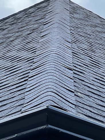 A close-up view of a roof with dark, overlapping shingles creating a textured surface. The shingles appear to be evenly laid with a symmetrical design.