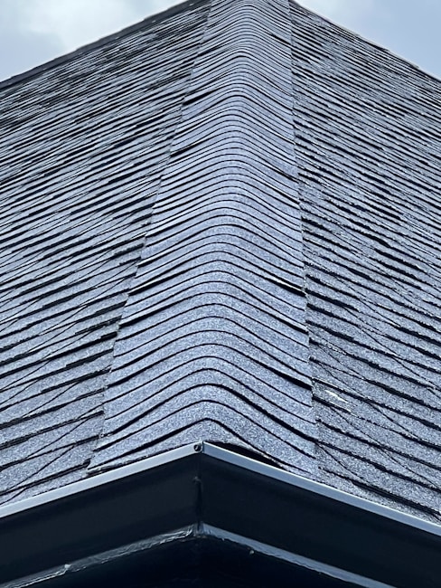 A close-up view of a roof with dark, overlapping shingles creating a textured surface. The shingles appear to be evenly laid with a symmetrical design.