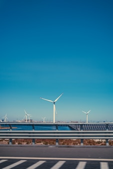 Several wind turbines stand tall in an open landscape under a clear blue sky. In the foreground, a road with metal guardrails is visible, indicating motion or travel. The background includes a body of water and a distant shore with buildings or structures. The scene suggests a blend of nature and technology.