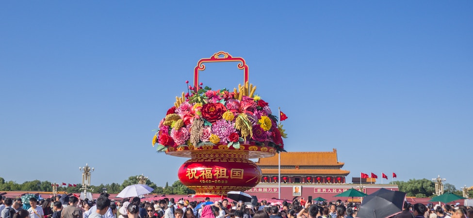 A large, decorative floral arrangement sits prominently against a clear blue sky. Brightly colored flowers, including pinks, reds, and yellows, are arranged in an ornate basket with red and gold designs. Below, a crowd of people gathers, some holding umbrellas, in front of a traditional building with a tiled roof and Chinese flags fluttering.