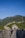 A group of people walk along the iconic stone path of the Great Wall of China, surrounded by lush green hills under a clear blue sky. The winding structure stretches into the distance, with a small gatehouse visible in the foreground.