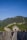 A group of people walk along the iconic stone path of the Great Wall of China, surrounded by lush green hills under a clear blue sky. The winding structure stretches into the distance, with a small gatehouse visible in the foreground.