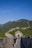 A group of people walk along the iconic stone path of the Great Wall of China, surrounded by lush green hills under a clear blue sky. The winding structure stretches into the distance, with a small gatehouse visible in the foreground.