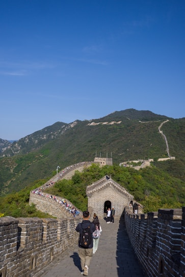 A group of people walk along the iconic stone path of the Great Wall of China, surrounded by lush green hills under a clear blue sky. The winding structure stretches into the distance, with a small gatehouse visible in the foreground.