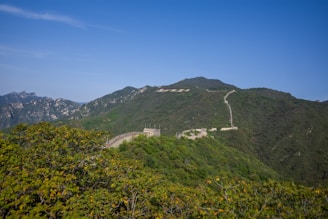 A beautiful landscape featuring a long stone wall snaking over lush green hills. The scene is set against a clear blue sky with distant mountain peaks visible on the left. The stone structure meanders smoothly along the ridge, highlighting the natural contours of the landscape.