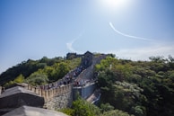 The scene showcases a section of the Great Wall of China winding through lush green hills under a clear blue sky. A large crowd of people is visible traversing the stone structure, highlighting its popularity as a tourist destination. Sunlight brightens the landscape, casting shadows on the wall and surrounding foliage.