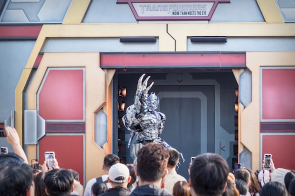 A metallic robot character poses dramatically at the entrance of a themed building, surrounded by a group of onlookers taking photos with their phones. The structure has a futuristic design with panels and an emblem at the top. The scene creates an engaging interaction between the character and the crowd.