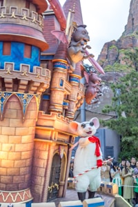 A whimsical scene featuring a castle structure with colorful and ornate decorations, including playful gargoyle-like creatures perched on the towers. In front of the castle, a large mouse character wearing sunglasses and a red scarf is holding a candy cane. A group of people is gathered nearby, enjoying the festive atmosphere.
