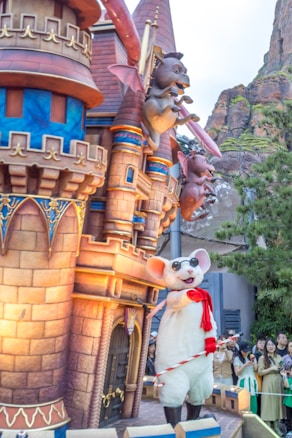 A whimsical scene featuring a castle structure with colorful and ornate decorations, including playful gargoyle-like creatures perched on the towers. In front of the castle, a large mouse character wearing sunglasses and a red scarf is holding a candy cane. A group of people is gathered nearby, enjoying the festive atmosphere.