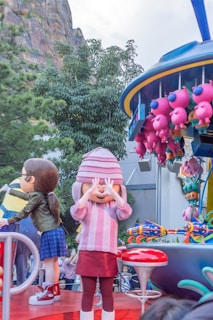 A lively parade scene featuring large, colorful cartoon character balloons and floats. One character is wearing a striped pink sweater and matching hat while covering their eyes. Another character beside them has brown hair and is wearing a green jacket and blue plaid skirt. A group of pink, pig-like balloons are attached to a nearby float. The background includes greenery and rock formations.
