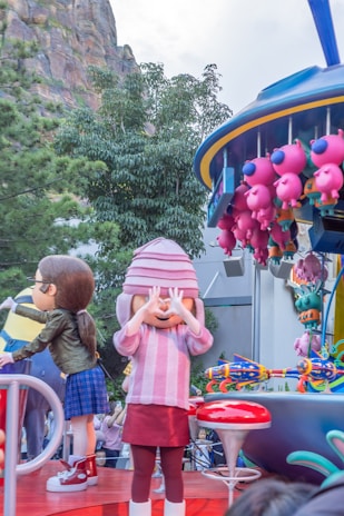 A lively parade scene featuring large, colorful cartoon character balloons and floats. One character is wearing a striped pink sweater and matching hat while covering their eyes. Another character beside them has brown hair and is wearing a green jacket and blue plaid skirt. A group of pink, pig-like balloons are attached to a nearby float. The background includes greenery and rock formations.