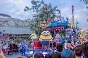 A lively parade scene features a float decked with colorful decorations and characters. The central characters appear to be yellow, cylindrical creatures with goggles, engaging with the crowd. A large crowd of onlookers, including excited children and adults, captures the moment with their phones. In the background, there is a building with artistic wall designs and windows.