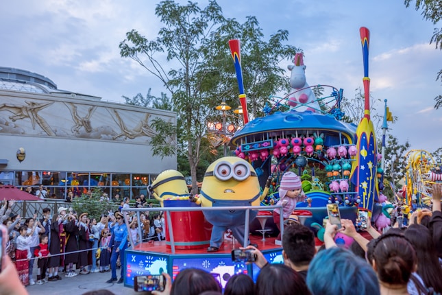 A lively parade scene features a float decked with colorful decorations and characters. The central characters appear to be yellow, cylindrical creatures with goggles, engaging with the crowd. A large crowd of onlookers, including excited children and adults, captures the moment with their phones. In the background, there is a building with artistic wall designs and windows.