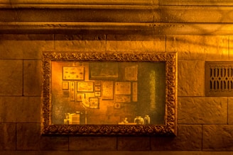 A framed painting hangs on a stone wall, illuminated by warm, amber lighting. The painting features various framed pictures and scientific illustrations arranged in a collage-like manner. Above the painting is a detailed, ornate molding, and to the right, a decorative vent is carved into the stone.