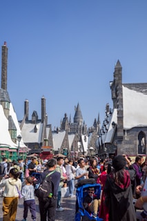 A bustling outdoor scene with a large crowd of people walking through a themed village area, featuring quaint, snow-topped roofs and stone buildings with spires reminiscent of a medieval or fantasy setting. Bright, clear blue skies provide a backdrop to the scene.