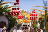 A festive outdoor area features many vibrant red lanterns hanging from wooden arches. In the background, a large decorative sign depicts a cartoon panda in an action pose, surrounded by colorful fireworks imagery. A crowd of people walk underneath, some looking around at the decor.