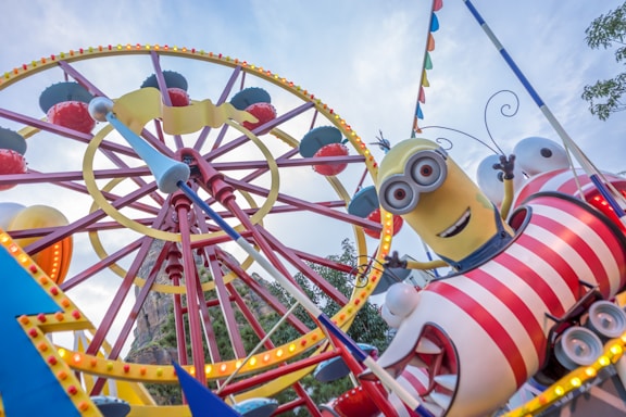 A whimsical amusement park ride with a large Ferris wheel in the background, featuring a cheerful character with yellow skin, goggles, and overalls riding on a bright red and white striped carriage. Colorful lights and flags decorate the ride, creating a festive atmosphere.