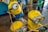 Three animated yellow characters with goggles and blue overalls are gathered together. One character has a single eye and is playfully sticking out of a popcorn bucket, while others are interacting playfully. The background includes a wooden floor and people lined up behind a rope barrier.