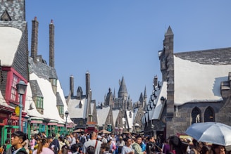 A bustling street filled with people set in a fantastical village with medieval-style architecture featuring pointed roofs and chimneys under a bright blue sky. Snow-like accents cover the roofs, and in the distance, a large castle-like structure is visible. Various shops, colorful awnings, and signs line the street, adding to the vibrant atmosphere.