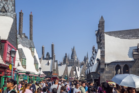 A bustling street filled with people set in a fantastical village with medieval-style architecture featuring pointed roofs and chimneys under a bright blue sky. Snow-like accents cover the roofs, and in the distance, a large castle-like structure is visible. Various shops, colorful awnings, and signs line the street, adding to the vibrant atmosphere.