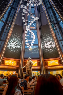 A large, metallic spiral structure resembling a DNA helix is suspended from the ceiling in a tall, futuristic hall. Two large illuminated signs with 'Jurassic World' logos are prominently displayed on either side of the room. Below the helix is a bronze statue of a man standing on a pedestal. A crowd of people is gathered at the bottom, creating a busy and bustling atmosphere.