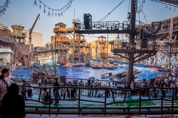An outdoor amphitheater with a large pool of water is set up for a live show. The stage is designed to resemble an industrial or post-apocalyptic world with elaborate metal structures, scaffolding, and decorative elements. The audience is composed of people of various ages, casually dressed, and seated or standing, eagerly anticipating the performance. Overhead, string lights add to the setting's ambiance.