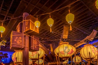 A warmly lit scene featuring ornate hanging lanterns with intricate designs. Some lanterns have spherical shapes, while others are box-shaped with patterns and images. Wooden beams and support structures are visible, contributing to a rustic ambiance.