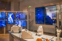 A high-tech laboratory environment featuring multiple digital screens displaying complex graphical data, including a large DNA helix. The room contains advanced laboratory equipment on a sleek table and a chair draped with a white lab coat. Soft lighting and a futuristic aesthetic contribute to the overall ambiance.