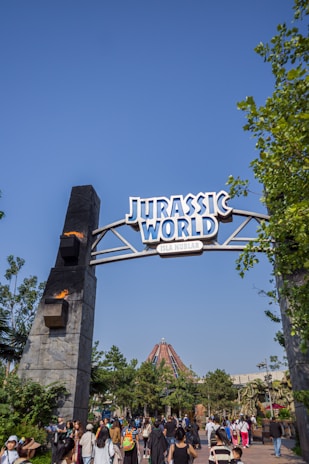 An archway with 'Jurassic World' written on it stands tall amidst a clear blue sky. Flames are visible on the side posts of the arch. Below, a crowd of people is gathered, with greenery and a large building structure in the background.