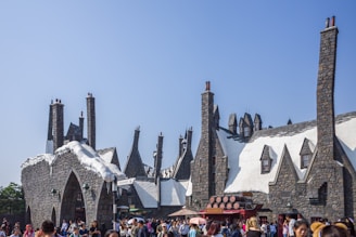 A bustling scene of a fictional village with snow-covered rooftops and numerous chimneys. The architecture features stone buildings that resemble a fantasy setting, with pointed roofs and narrow windows. A crowd of visitors strolls through the area, likely exploring a themed attraction.