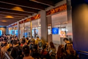 A crowded indoor space with a group of people in a queue. They appear to be waiting in line, facing a series of windows or counters on the right. Behind the windows are interior rooms with illuminated screens and displays, suggesting a technological or interactive exhibit.
