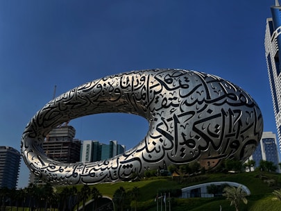 A striking, futuristic building featuring an elliptical shape with intricate Arabic calligraphy across its metallic surface, set against a backdrop of modern skyscrapers and a clear blue sky.