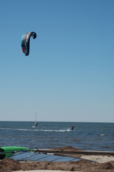 Two people are engaged in kitesurfing on the open sea. The sky is clear blue, and the kite is colorful, predominantly black with blue and red accents. In the foreground, a section of sandy beach with seaweed and a green kayak can be seen.