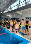 A group of people standing beside an indoor swimming pool, wearing swim caps and holding colorful pool noodles. The interior features large windows with natural light streaming in, a high ceiling with exposed beams, and a wall-mounted clock.