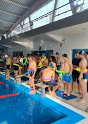 A group of people standing beside an indoor swimming pool, wearing swim caps and holding colorful pool noodles. The interior features large windows with natural light streaming in, a high ceiling with exposed beams, and a wall-mounted clock.