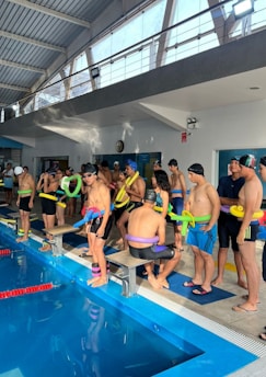 A group of people standing beside an indoor swimming pool, wearing swim caps and holding colorful pool noodles. The interior features large windows with natural light streaming in, a high ceiling with exposed beams, and a wall-mounted clock.