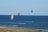 Three kitesurfers are seen gliding over the ocean with colorful kites flying high in the air. The sea stretches into the horizon, and the foreground displays a strip of grassy land.