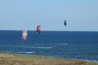 Three kitesurfers are seen gliding over the ocean with colorful kites flying high in the air. The sea stretches into the horizon, and the foreground displays a strip of grassy land.