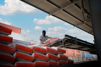 A person wearing a mask and cap is handling large bags of goods, stacking them on a conveyor belt under a metal roof. The sky is partly cloudy, and there are shipping containers in the background.