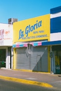A storefront with a large bright yellow sign that reads 'La Gloria' with additional text indicating it sells clothing for babies, baptisms, and ceremonies. The shop has a closed metal shutter with a striped awning in shades of pink and blue.