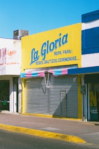 A storefront with a large bright yellow sign that reads 'La Gloria' with additional text indicating it sells clothing for babies, baptisms, and ceremonies. The shop has a closed metal shutter with a striped awning in shades of pink and blue.
