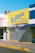 A storefront with a large bright yellow sign that reads 'La Gloria' with additional text indicating it sells clothing for babies, baptisms, and ceremonies. The shop has a closed metal shutter with a striped awning in shades of pink and blue.