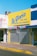 A storefront with a large bright yellow sign that reads 'La Gloria' with additional text indicating it sells clothing for babies, baptisms, and ceremonies. The shop has a closed metal shutter with a striped awning in shades of pink and blue.