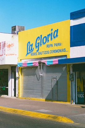 A storefront with a large bright yellow sign that reads 'La Gloria' with additional text indicating it sells clothing for babies, baptisms, and ceremonies. The shop has a closed metal shutter with a striped awning in shades of pink and blue.