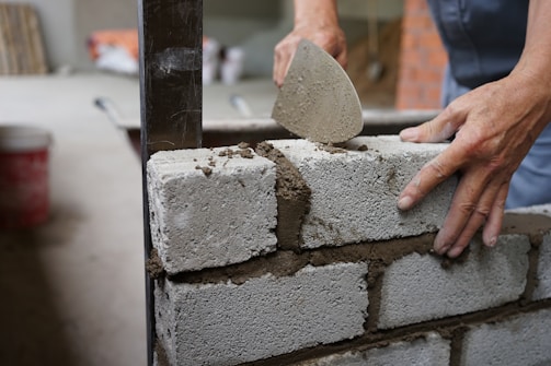 A person is using a trowel to apply mortar between cinder blocks while constructing a wall. The setting appears to be a construction site with additional building materials visible in the background.