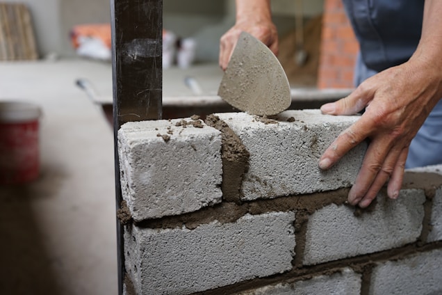 A person is using a trowel to apply mortar between cinder blocks while constructing a wall. The setting appears to be a construction site with additional building materials visible in the background.