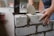 A person is using a trowel to apply mortar between cinder blocks while constructing a wall. The setting appears to be a construction site with additional building materials visible in the background.