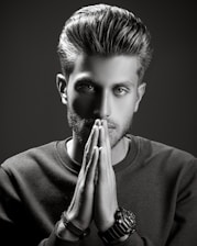 A grayscale portrait of a man with styled hair, looking intently at the camera with his hands placed together in front of his face. He wears a textured sweater and several bracelets on one wrist. The lighting is dramatic, creating strong shadows and highlights to emphasize his facial features.