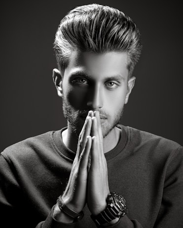 A grayscale portrait of a man with styled hair, looking intently at the camera with his hands placed together in front of his face. He wears a textured sweater and several bracelets on one wrist. The lighting is dramatic, creating strong shadows and highlights to emphasize his facial features.