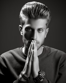 A grayscale portrait of a man with styled hair, looking intently at the camera with his hands placed together in front of his face. He wears a textured sweater and several bracelets on one wrist. The lighting is dramatic, creating strong shadows and highlights to emphasize his facial features.